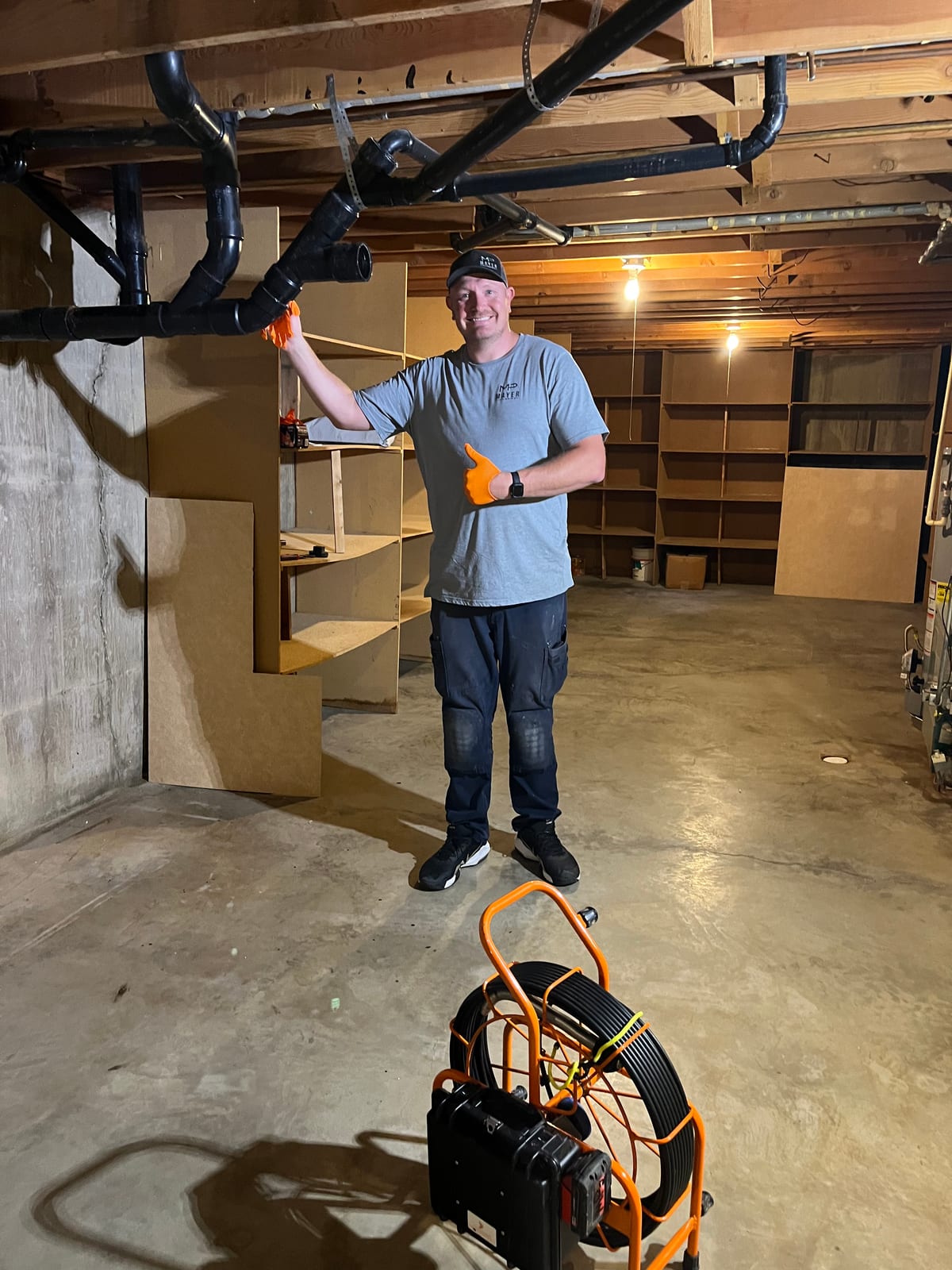 Mayer Plumbing technician beside basement drain piping and a drain camera reel
